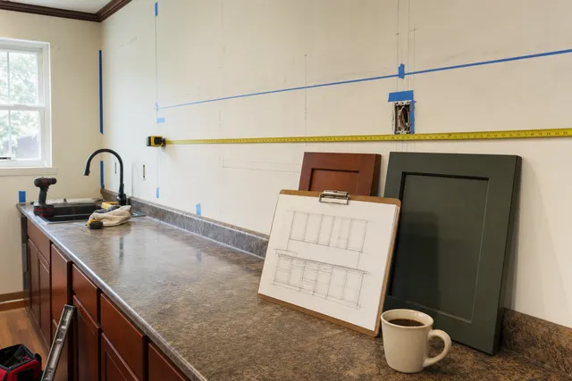 Tape measure and blue tape laid out on a bare kitchen wall next to cherry and green door samples and a sketched elevation
